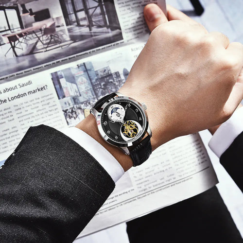 Hand wearing a black wristwatch with a detailed face, reading a newspaper in the background.