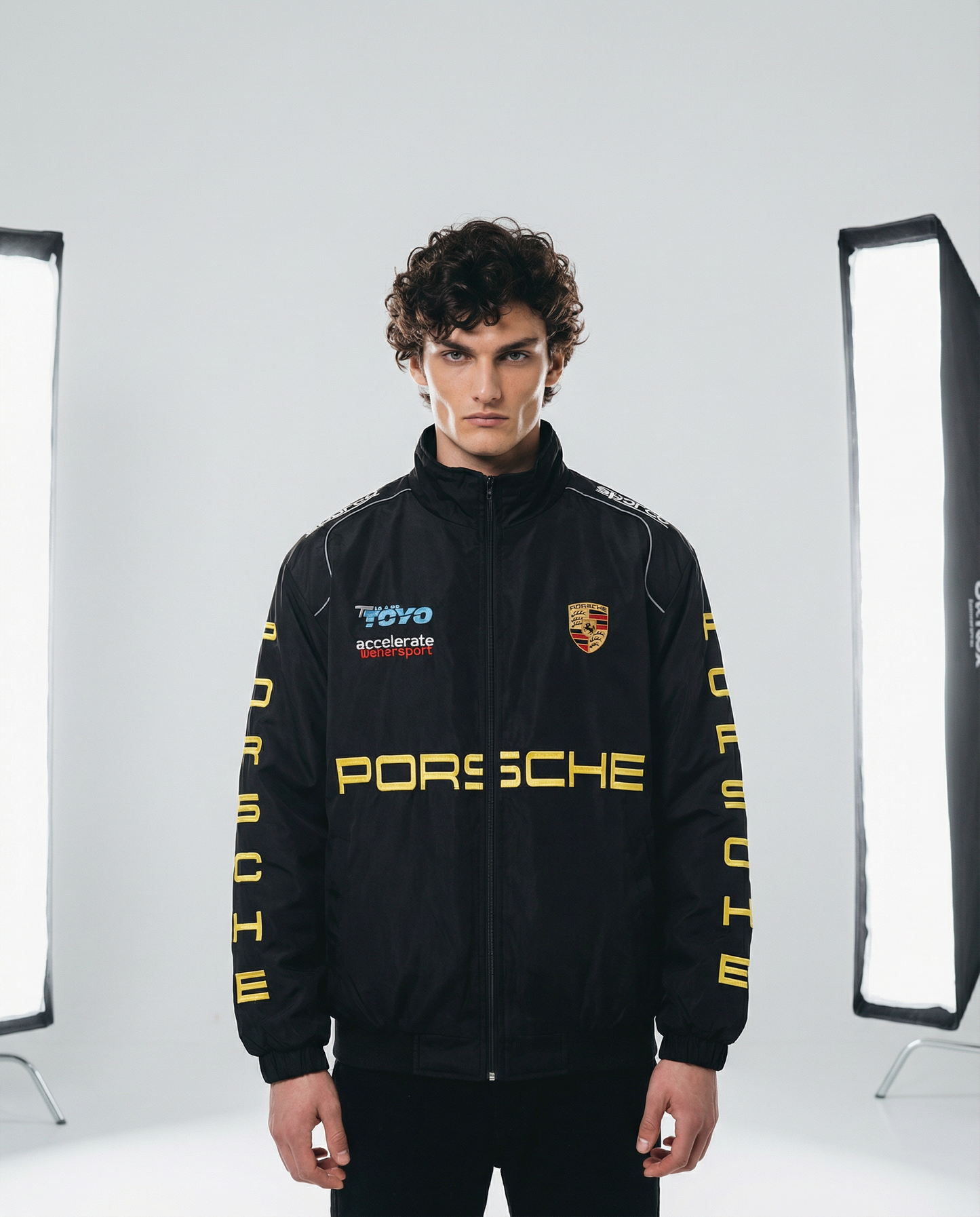 Person wearing a black Porsche jacket with logos in a studio setting. Front view of Porsche Racing Jacket showing relaxed fit, full zip closure, and bold branding
