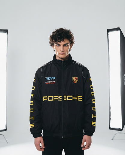 Person wearing a black Porsche jacket with logos in a studio setting. Front view of Porsche Racing Jacket showing relaxed fit, full zip closure, and bold branding
