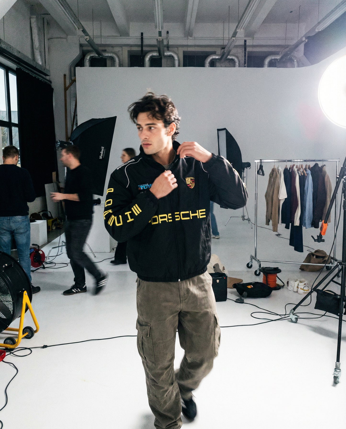 Man wearing a Porsche jacket in a studio setting with equipment and clothing racks. Behind-the-scenes studio photoshoot featuring Porsche Racing Jacket worn by models
