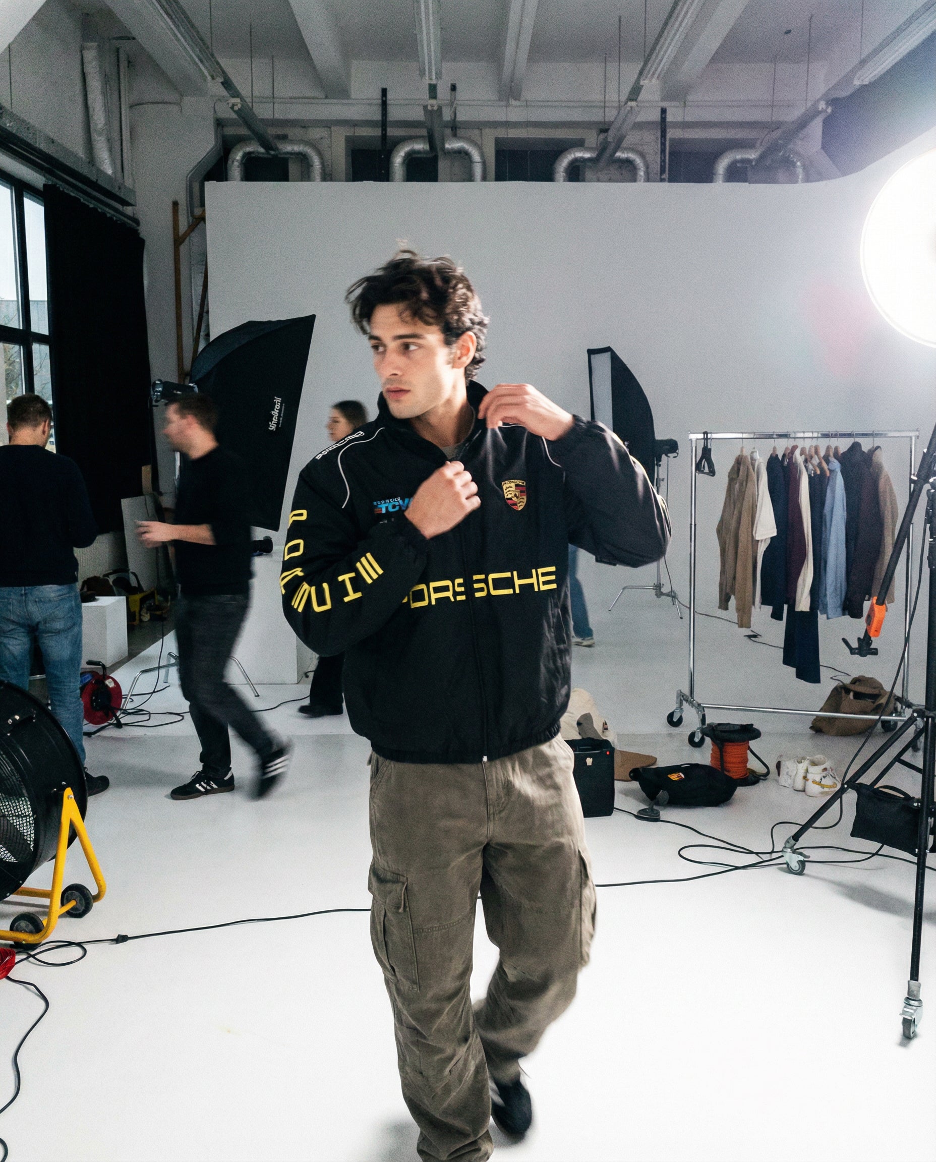 Man wearing a Porsche jacket in a studio setting with equipment and clothing racks. Behind-the-scenes studio photoshoot featuring Porsche Racing Jacket worn by models
