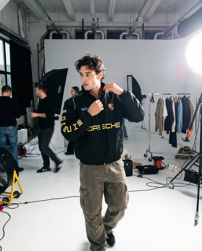 Man wearing a Porsche jacket in a studio setting with equipment and clothing racks. Behind-the-scenes studio photoshoot featuring Porsche Racing Jacket worn by models

