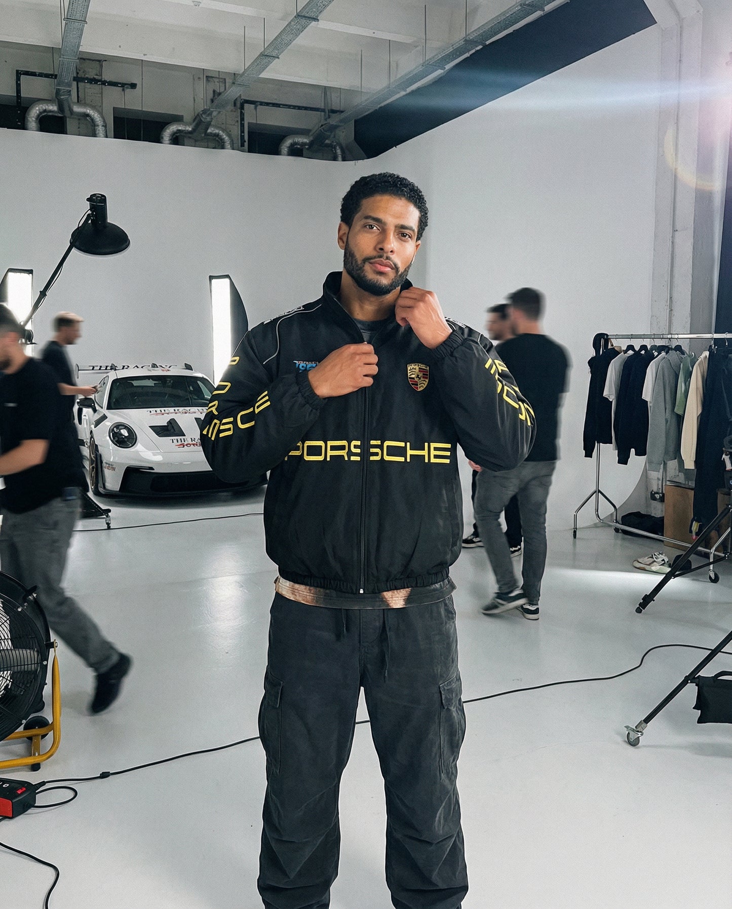 Person wearing a Porsche jacket in a studio setting with a car in the background. Porsche Racing Jacket styled for luxury motorsport streetwear in studio setting
